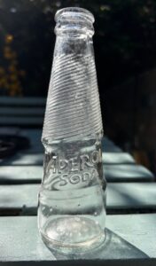 A small glass bottle with Aperol Soda imprinted in the glass sits on a green table.